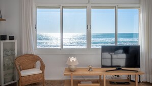 Ett rum med ett stort fönster med utsikt över havet, en tv på ett träställ, en flätad fåtölj och ett glasskåp.