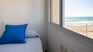 En säng med en blå kudde och ett randigt överkast, ett fönster med utsikt över stranden och en ljusströmbrytare på väggen.