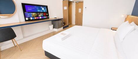Una habitación de hotel con una cama, un escritorio con un monitor de computadora, una silla y un refrigerador pequeño.