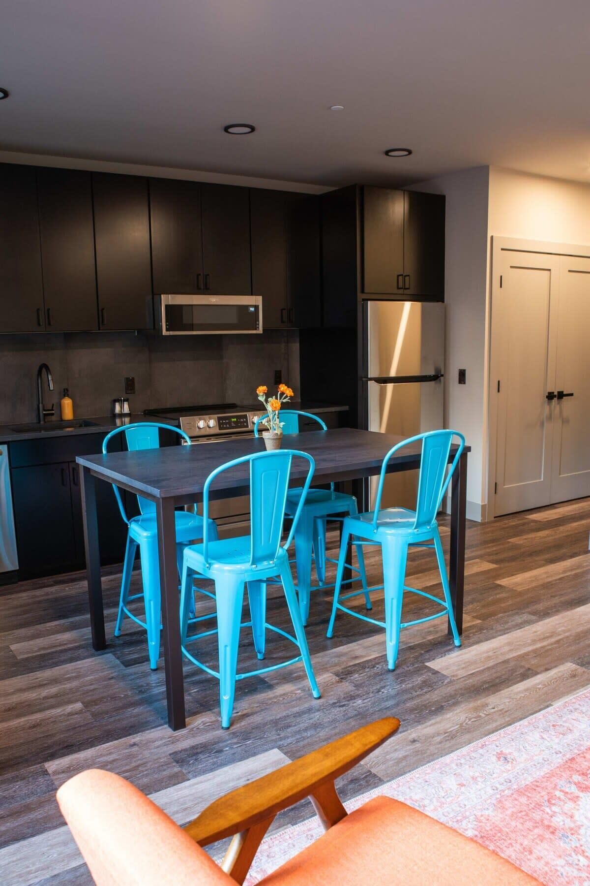 A modern kitchen with dark cabinets, a stainless steel refrigerator, and a microwave. There is a dining table with four blue chairs and a bench.