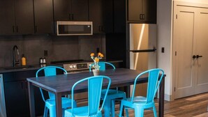 A modern kitchen with dark cabinets, a stainless steel refrigerator, and a microwave. There is a dining table with four blue chairs and a bench. - Modern State Unit 2 (Traverse City)