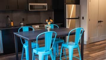 A modern kitchen with dark cabinets, a stainless steel refrigerator, and a microwave. There is a dining table with four blue chairs and a bench.