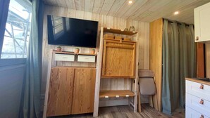 A compact room with a wooden ceiling, a flat-screen TV mounted on the wall, a small wooden cabinet with drawers, and a window with curtains.