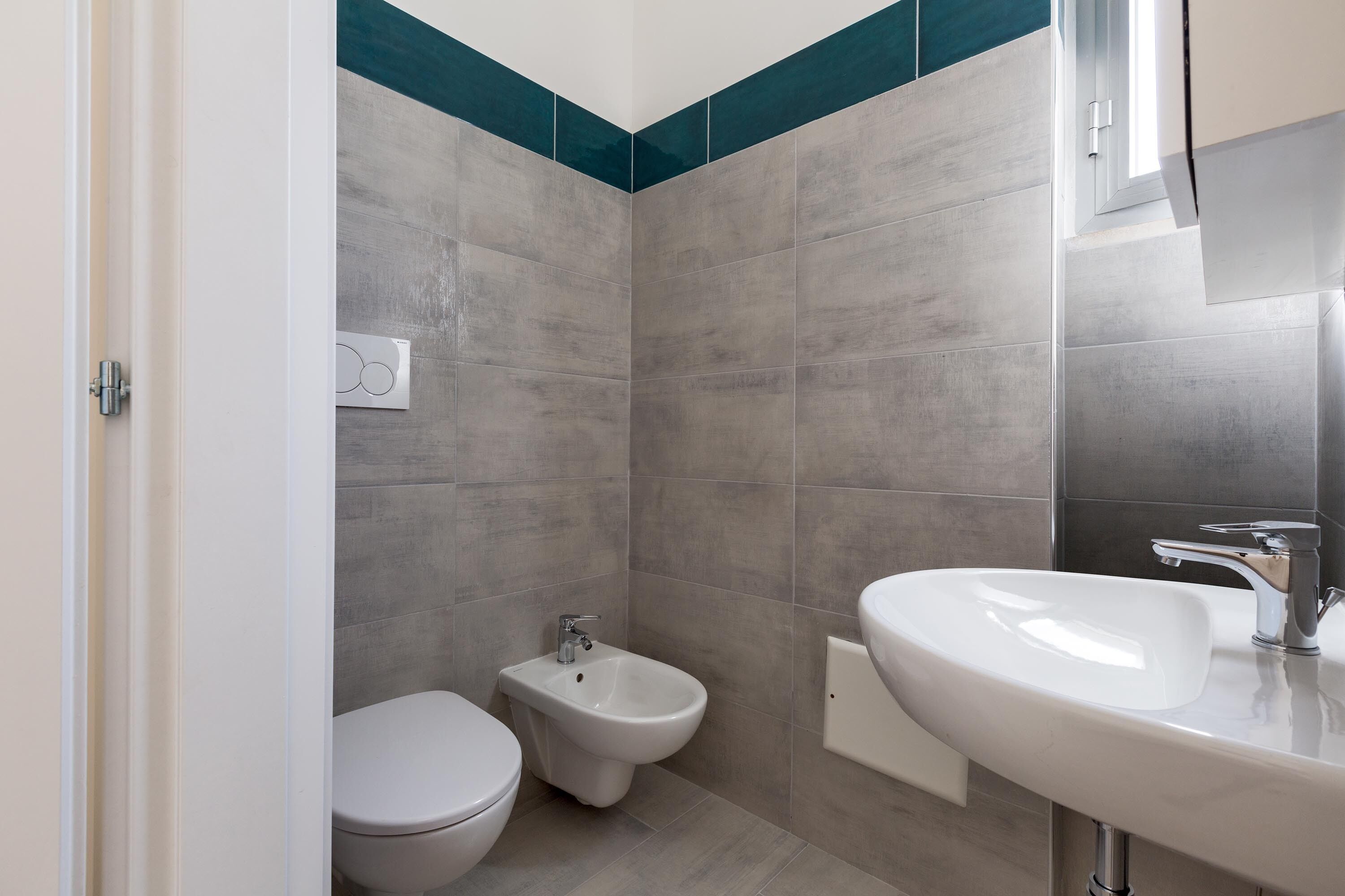 Un baño moderno con azulejos grises, un lavamanos blanco, un bidé y un inodoro.