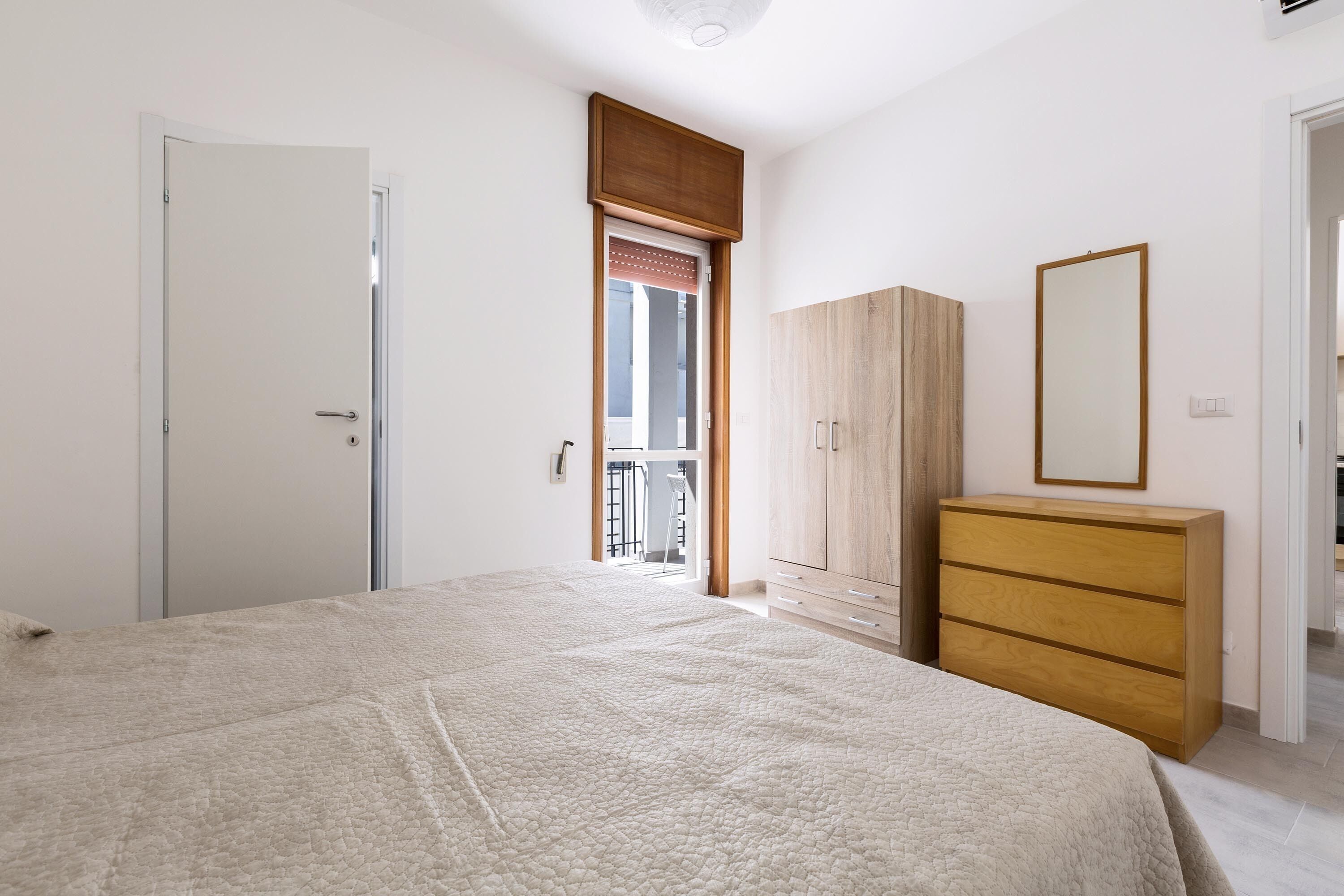 A bedroom with a bed, a wooden wardrobe, a dresser, and a door leading to a balcony.