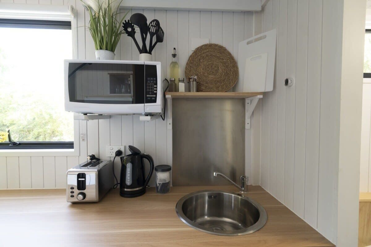 A compact kitchen with a microwave, toaster, and stainless steel sink. A potted plant and kitchen utensils are on the wall, and a woven basket is on the counter.