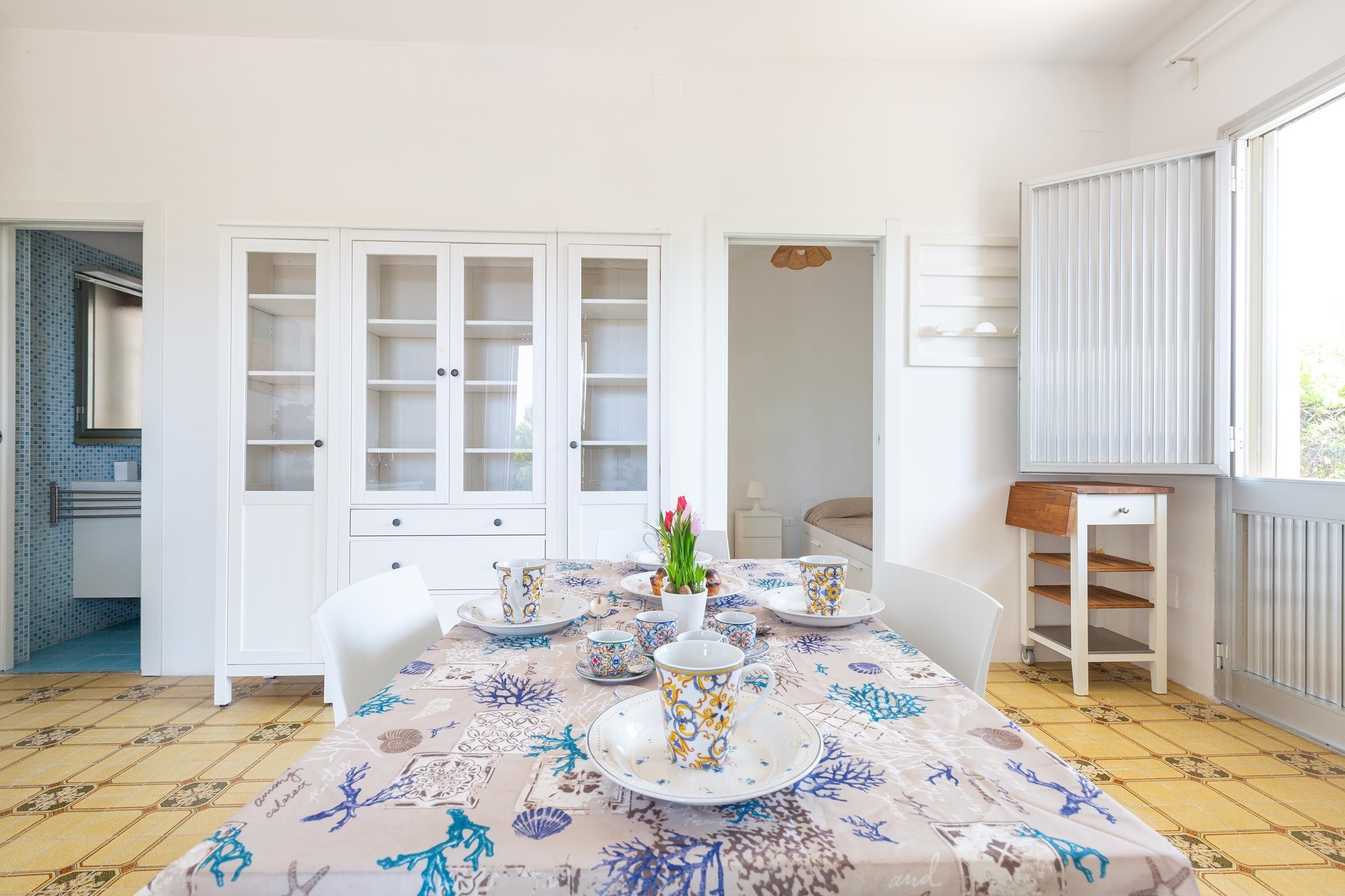 Un comedor con una mesa preparada para cuatro personas, armarios blancos, una ventana con persianas y un radiador.