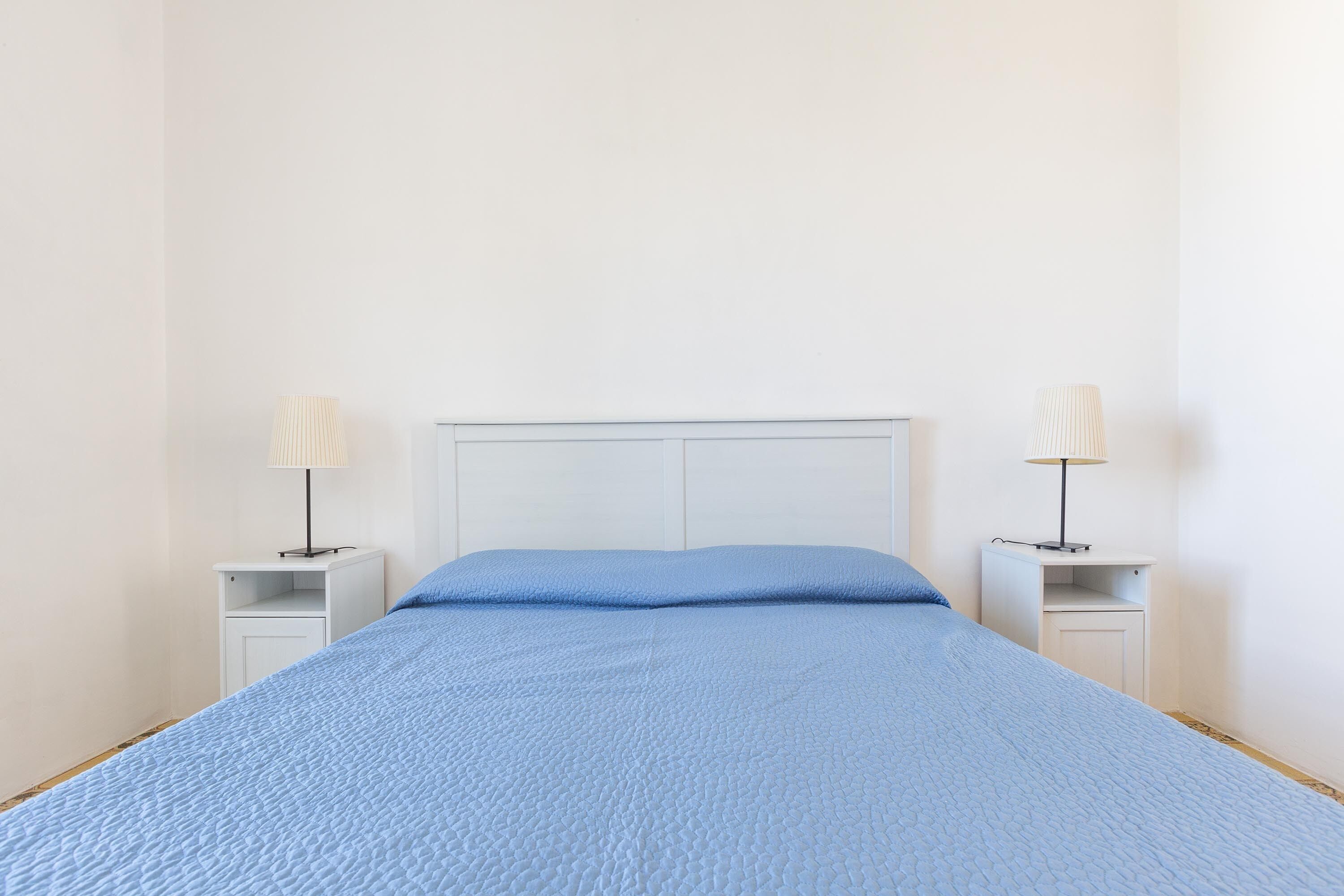 Een bed met een blauwe quilt, twee nachtkastjes met lampen en een wit hoofdeinde.
