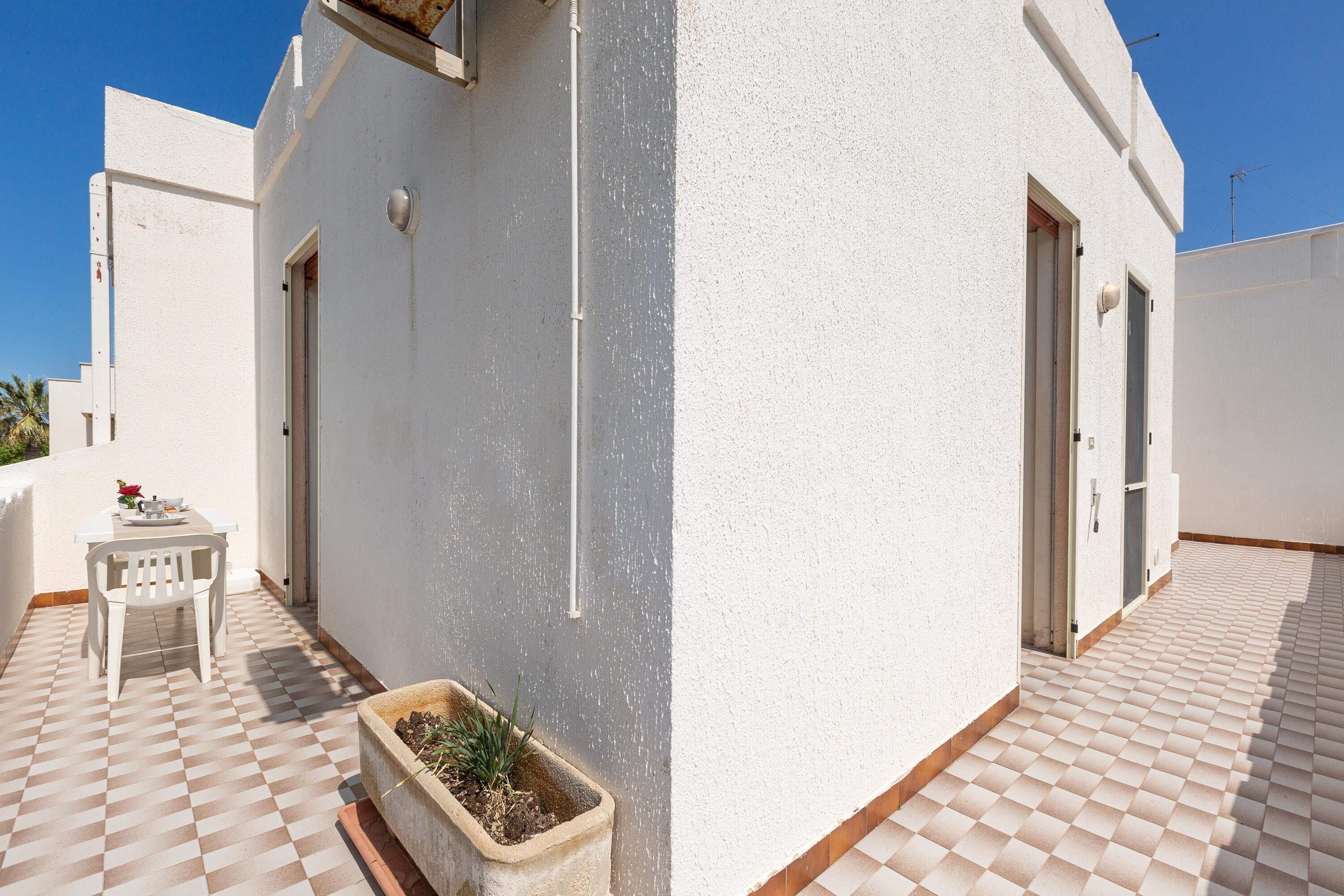 A white building with a tiled patio, a small table with chairs, and a planter with plants.