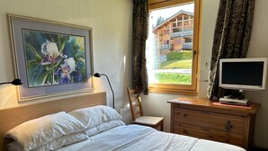 A bedroom with a bed, a desk with a computer, a painting, and a view of a building outside.