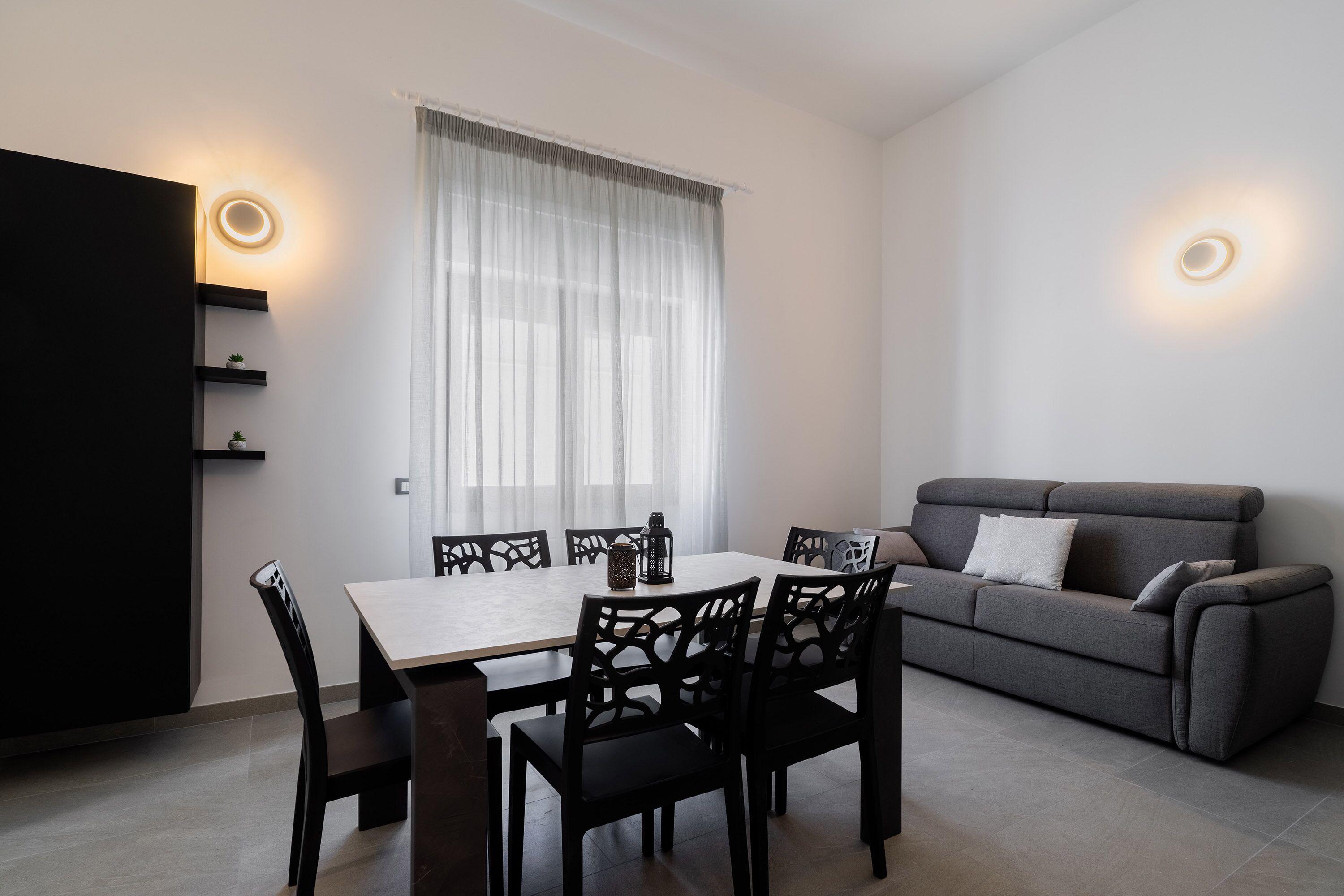 A modern living room with a dining area, featuring a sofa, a dining table with chairs, and a large window with sheer curtains.