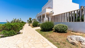 A pathway leading to a modern building with a white facade, stone paving, and surrounding greenery. - 3429 Residence Medusa - Suite Zeus PT (Porto Cesareo)