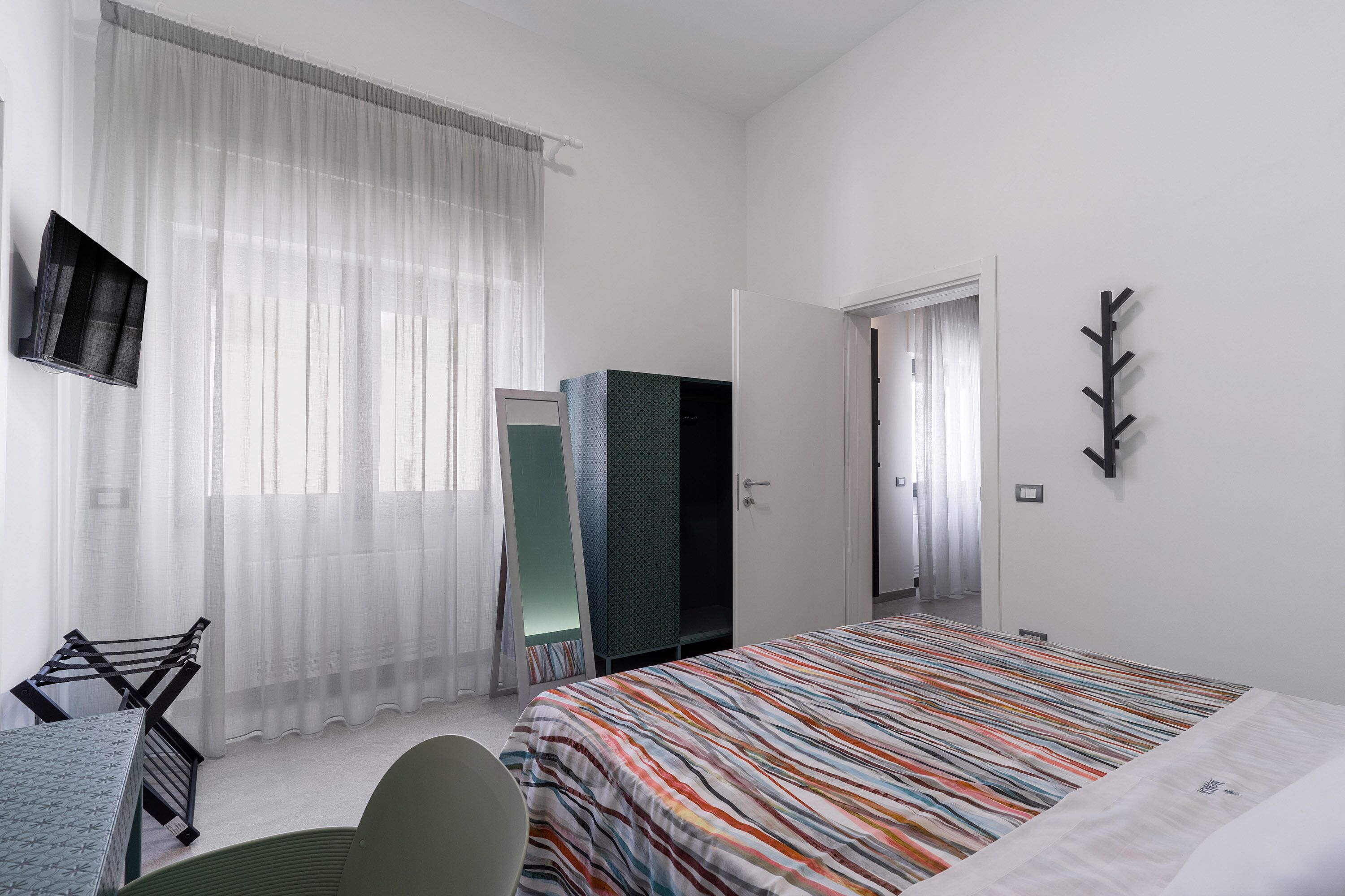 Habitación de hotel con una cama, televisión montada en la pared, armario y perchero.