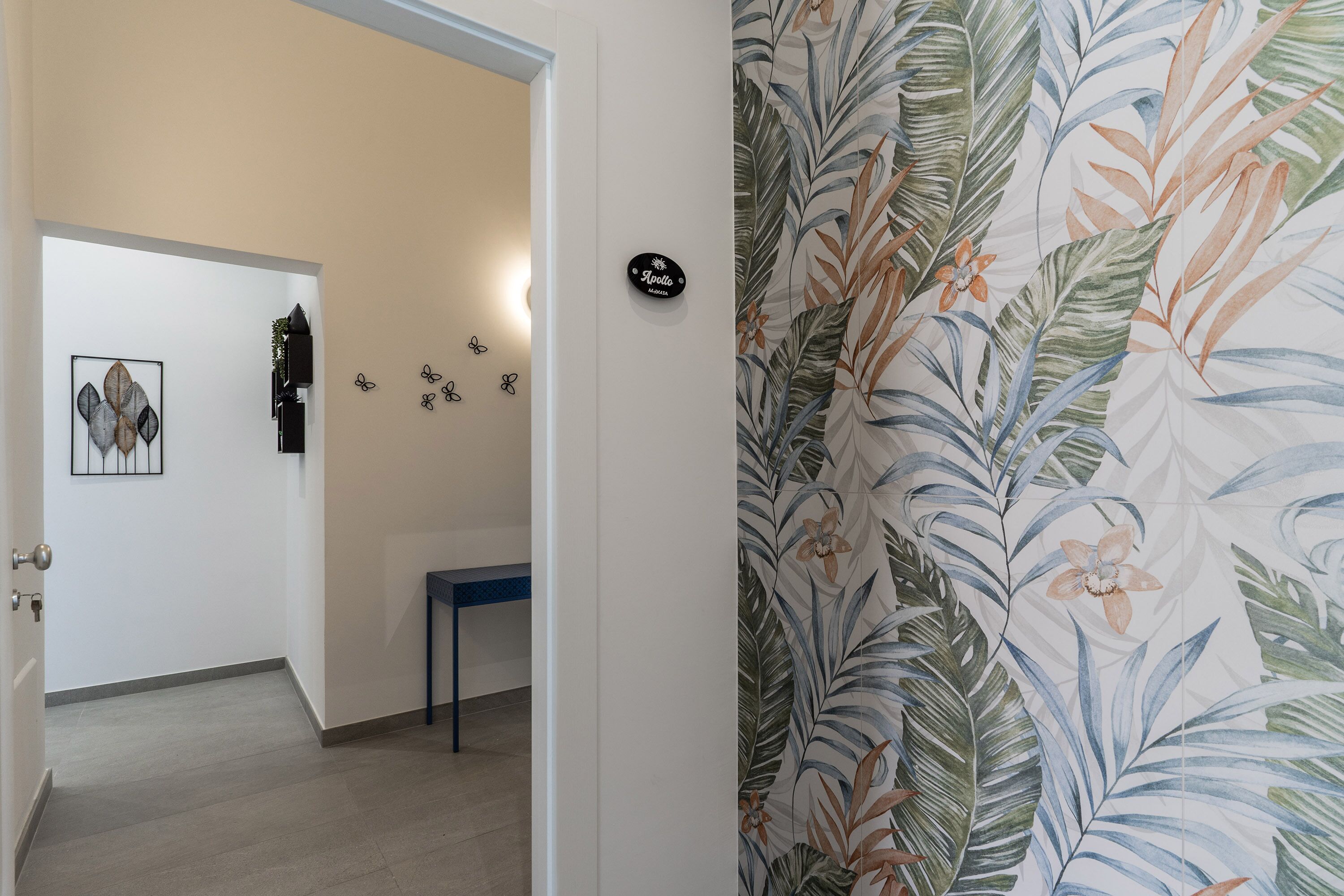 Ein Korridor mit einer Tür zu einem Zimmer, einem kleinen Tisch und einer Wand mit einem tropischen Blattmuster.