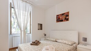 Ein Schlafzimmer mit einem großen Bett, einem Nachttisch, einem Fenster mit Vorhängen und einem an der Wand befestigten Kunstwerk.