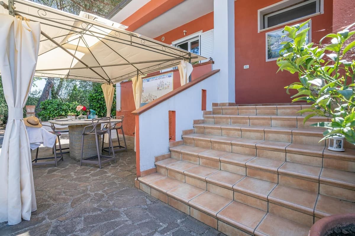 Un patio con mesa y sillas, un área cubierta con toldo blanco y una escalera de piedra que conduce a un edificio rojo.