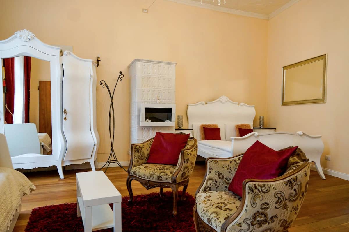 A bedroom with a bed, two armchairs, a white side table, a mirror, and a television.