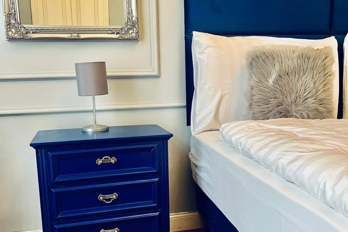 A bedroom with a dark blue nightstand, a lamp, a bed with white bedding, and a large mirror above the dresser.