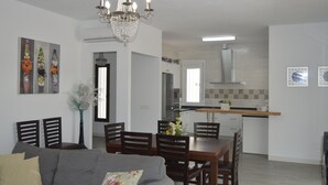 A modern living room with a grey sofa, wooden dining table, and chairs. There is a chandelier above the table and a kitchen area in the background. - Villa Ana, Chalet Independiente (AN)