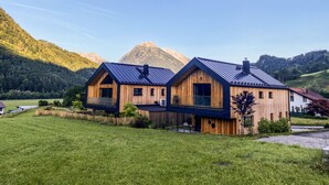 Ein modernes Holzhaus mit geneigtem Dach, umgeben von einer grünen Wiese und Bergen im Hintergrund.