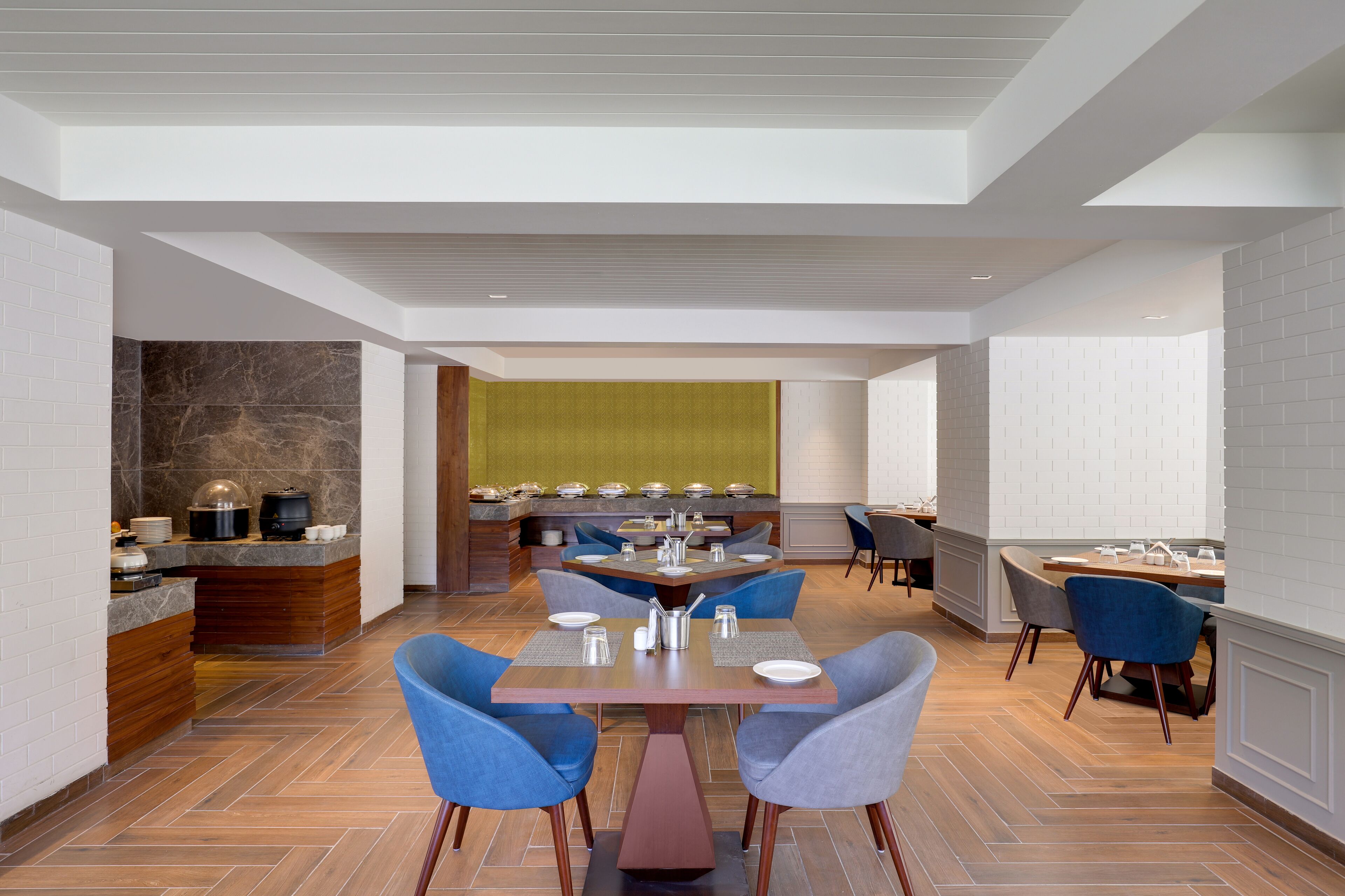 A modern dining area with a central table surrounded by chairs, a kitchen in the background, and a bright, well-lit space with a herringbone floor pattern.