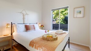 Ein ordentlich bezogenes Bett mit weißen Leinentüchern und einer pfirsichfarbenen Tagesdecke, ein Nachttisch mit einer Lampe, ein Fenster mit Blick ins Grüne und ein gerahmtes Bild an der Wand.