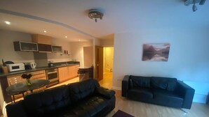 A modern living room with a black leather sofa, a dining table with a vase of flowers, and a kitchen area with wooden cabinets and a microwave. - Immaculate 1-bed Apartment in Birmingham (Birmingham)