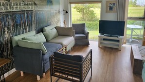 Ein modernes Wohnzimmer mit einem Ecksofa, einem kleinen Sessel, einer TV-Konsole und einem großen Fenster mit Blick ins Freie.