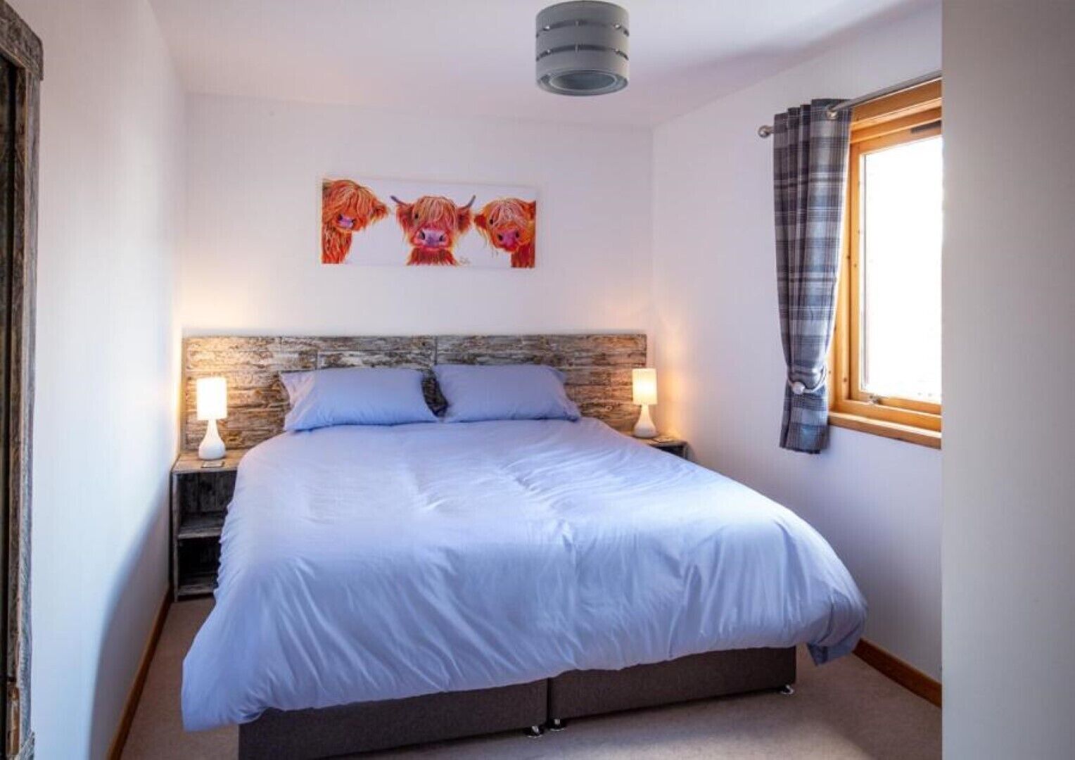 A bedroom with a large bed, two bedside tables with lamps, a headboard with a rustic texture, and a window with a plaid curtain.
