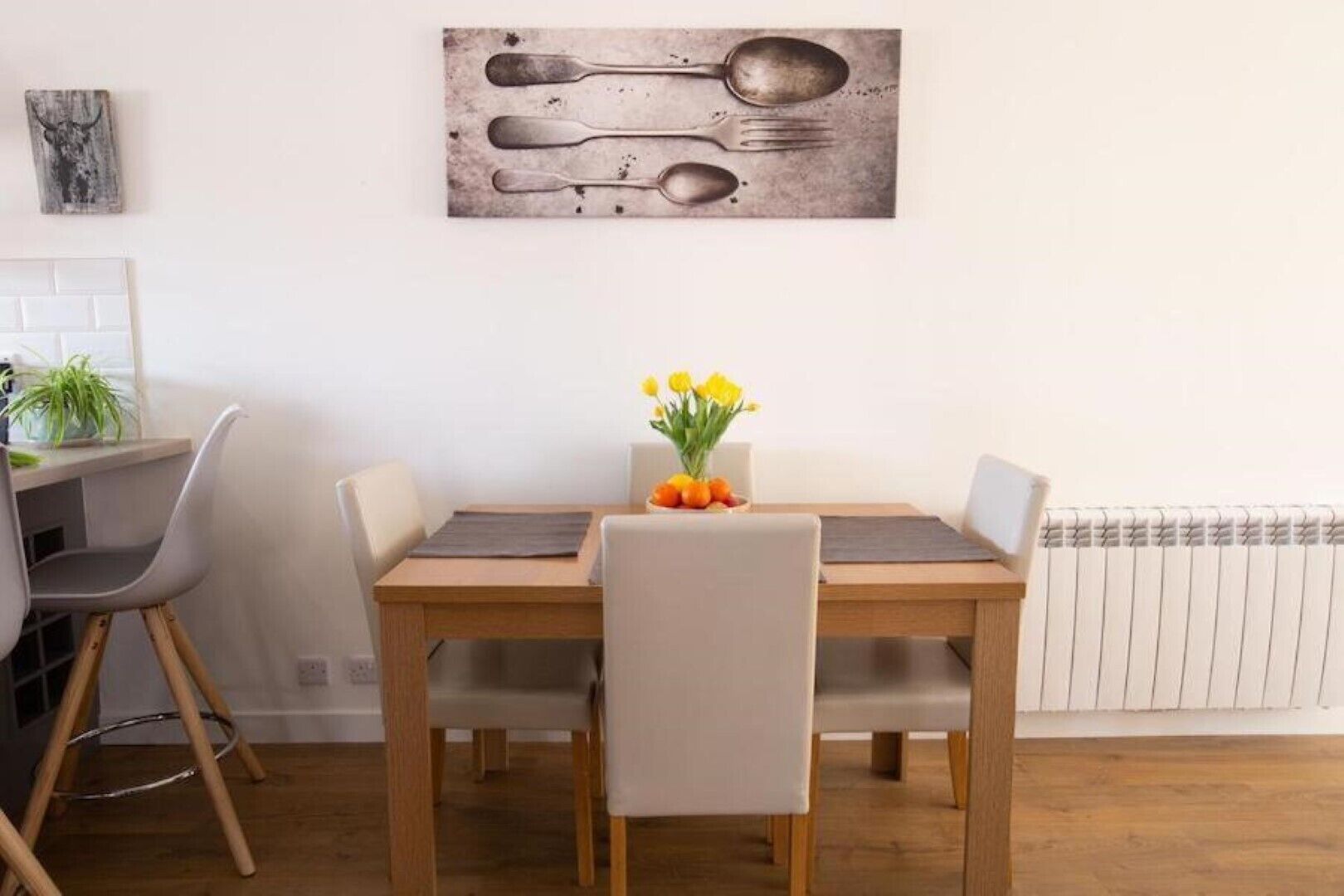 A dining area with a wooden table, four chairs, a vase with yellow flowers, and a wall-mounted artwork of utensils.