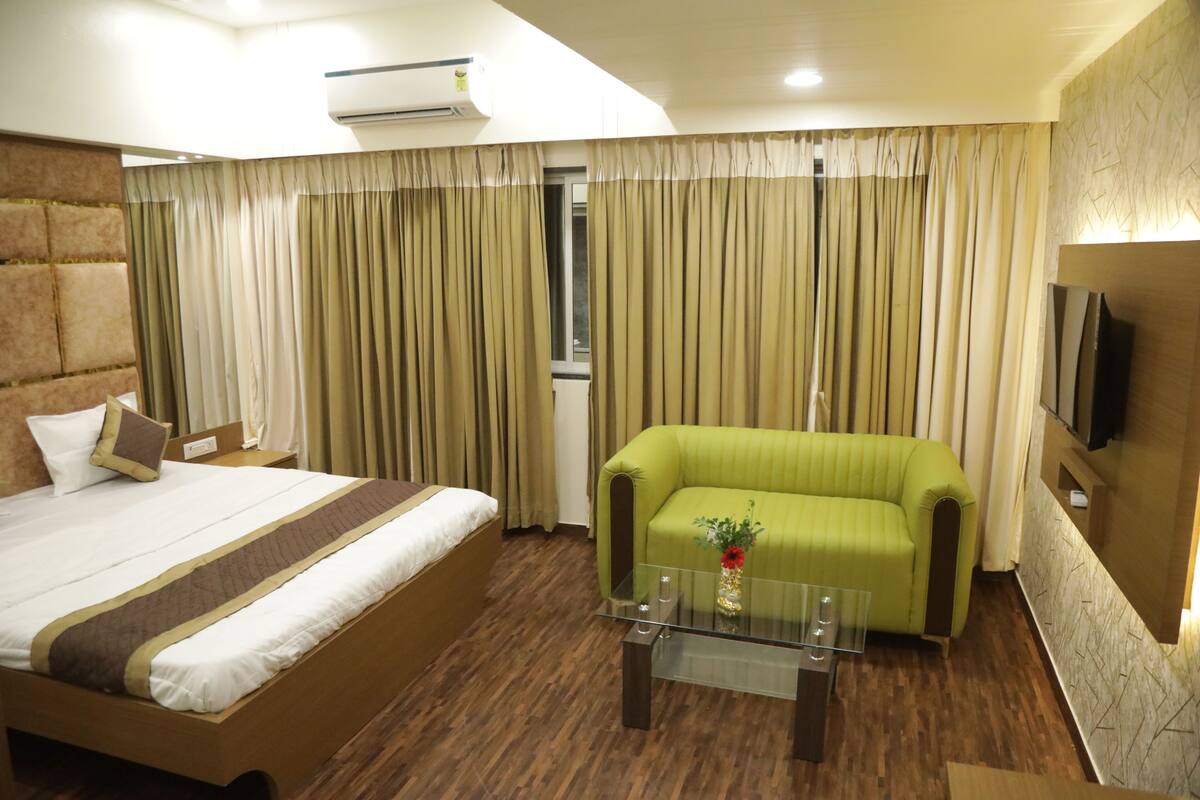 A hotel room with a bed, a green sofa, a glass coffee table, and a TV mounted on the wall.