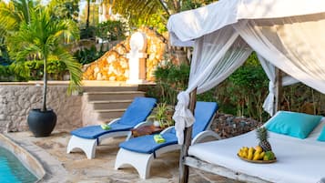 A poolside area with lounge chairs, a bed with a canopy, and a fruit basket.