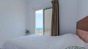 Een slaapkamer met een groot bed, uitzicht op zee door een schuifdeur en een balkon met gordijnen.