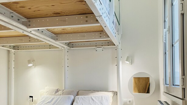 A bunk bed with white bedding, a circular wooden shelf, and a window with blinds.