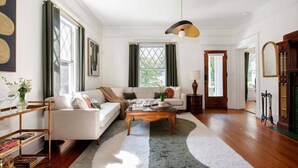 A spacious living room with a large sofa, a coffee table, and a rug. There is a window with curtains, a lamp, and a bookshelf.