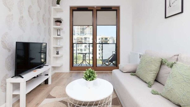 En moderne stue med et fladskærms-tv, et hvidt sofabord, en sofa med grønne puder og en stor skydedør, der fører ud til en balkon.