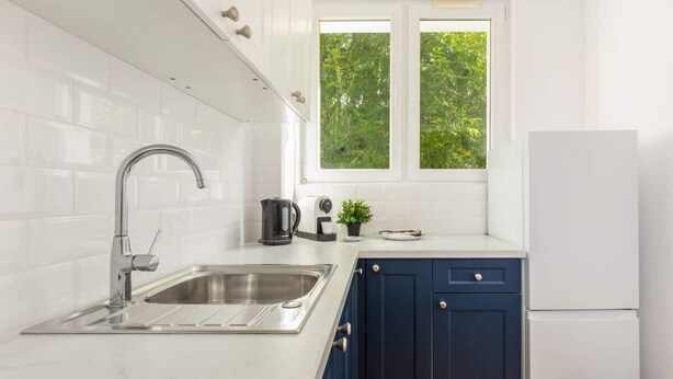 Une cuisine moderne avec des carreaux blancs, un évier en acier inoxydable, une fenêtre donnant sur un espace verdoyant et un réfrigérateur blanc.