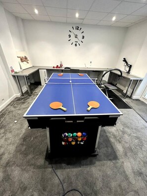 A ping pong table with paddles and balls, a clock, and a desk with a computer.