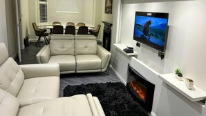 A modern living room with a fireplace, a mounted TV, a dining area with a table and chairs, and a sofa. - Serenity Haven 4-bed With Hottub,games,gym & More (Colchester)