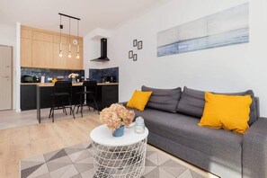 Un salon moderne avec un canapé gris, des coussins jaunes, une table basse blanche ornée d’un vase, et une cuisine en arrière-plan.