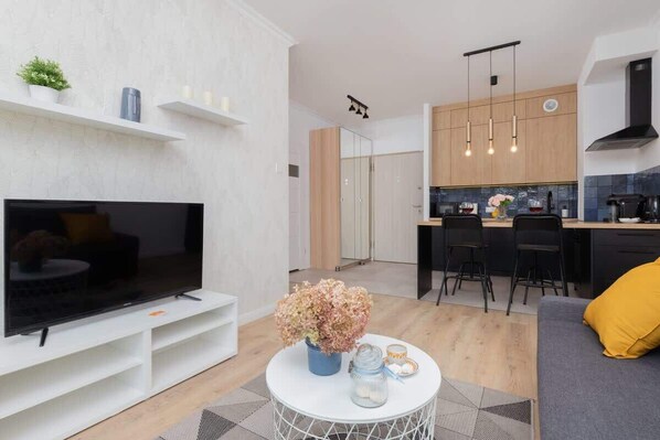 Un salon moderne avec un téléviseur à écran plat, une table basse agrémentée d’objets décoratifs et un canapé avec un coussin jaune. On aperçoit également la cuisine, avec ses meubles en bois et sa hotte noire.