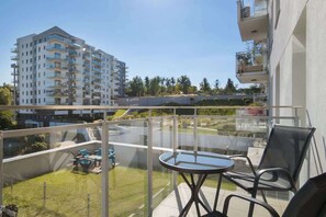 A balcony with a glass railing, a round table, and two chairs. In the background, there is a multi-story building and a grassy area with a fence.