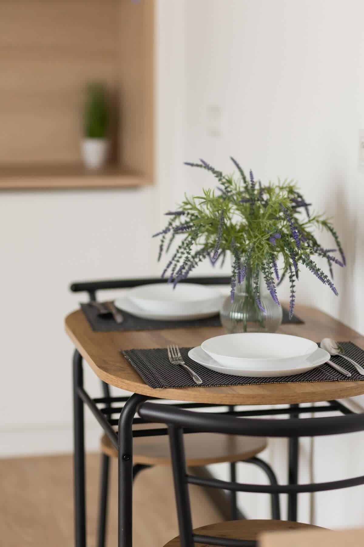 Una mesa de comedor con dos platos blancos, un florero de vidrio con follaje verde y un tenedor y una cuchara sobre una base para mantel negra.