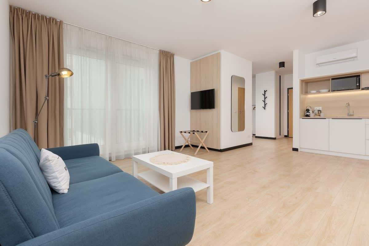 Una sala de estar moderna con un sofá azul, una mesa de centro blanca y una cocina con armarios blancos y un microondas.