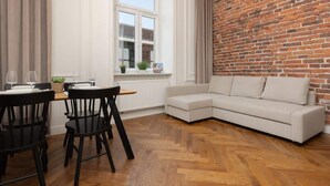 A modern living room with a white sofa, black chairs, a wooden herringbone floor, a brick wall, and a window with curtains.