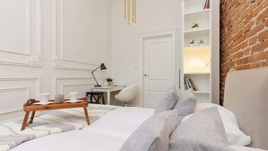 A neatly arranged bedroom with a bed, bedside table, lamp, and a shelf with books and plants.
