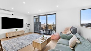 A modern living room with a flat-screen TV, a telescope, a wooden coffee table, a grey sofa, and a balcony with a view of the city. - Newly Built 3 Bedroom Townhouse with Ocean Views (Torquay)