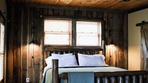 A wooden cabin room with a bed, two windows with blinds, and a wall-mounted light fixture.