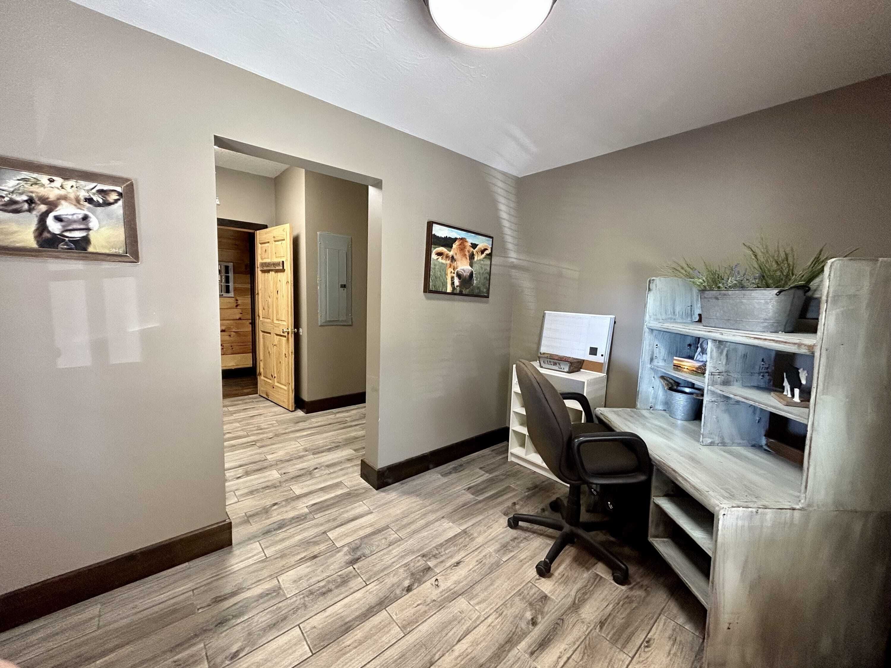 A modern office space with a wooden floor, a desk with a computer, a chair, and a bookshelf with plants.