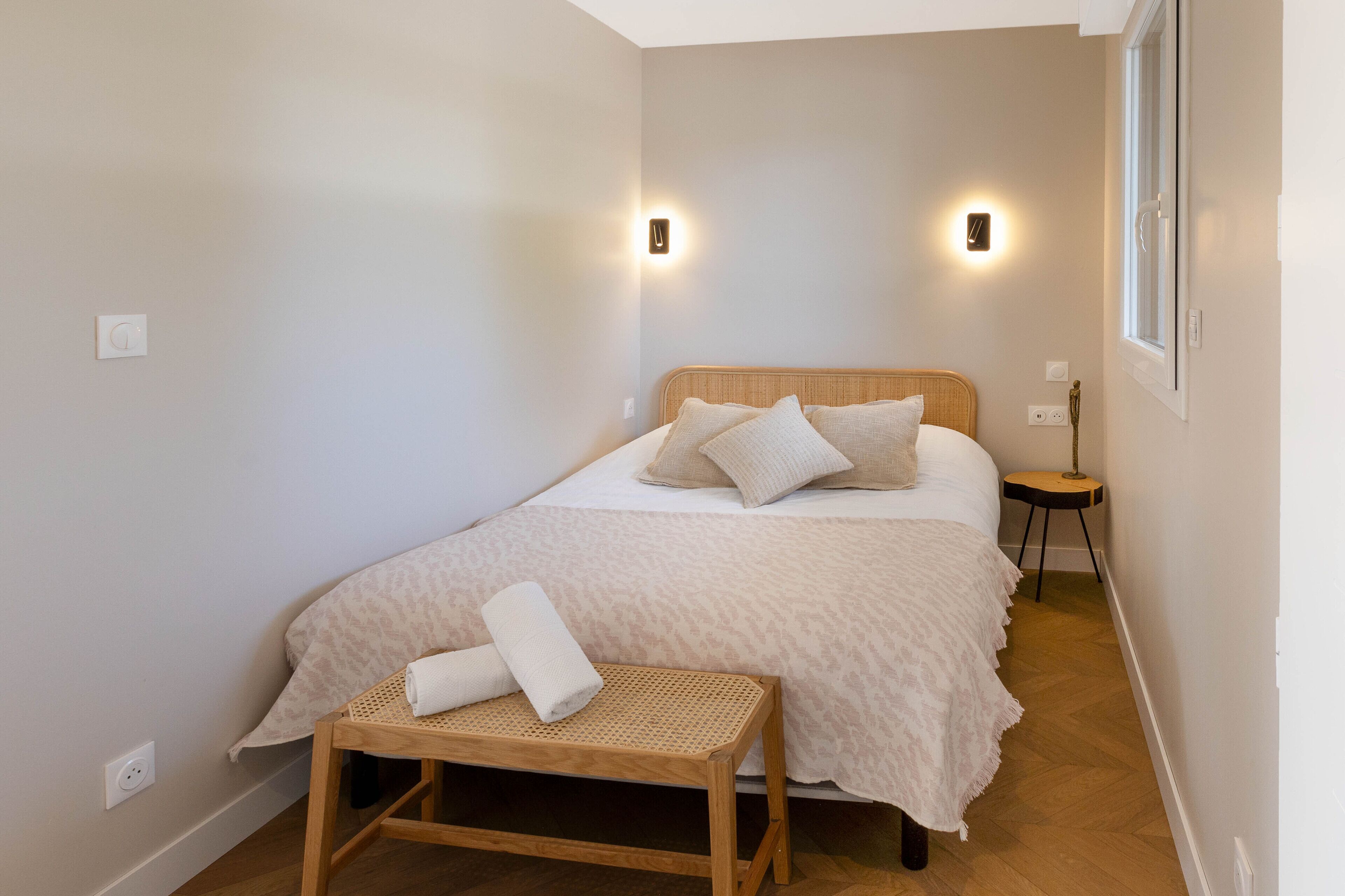 Un dormitorio con cama, lámparas de noche, una mesita y una ventana.